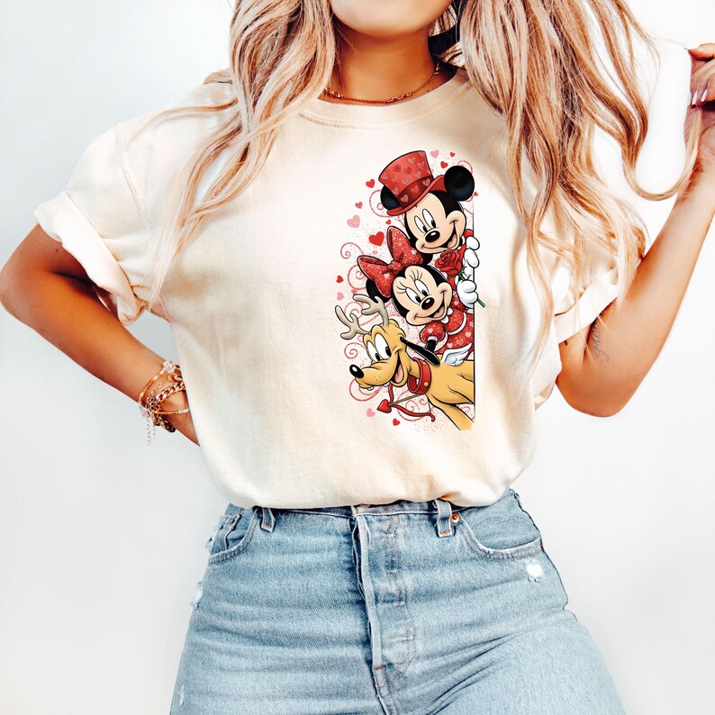 Puede incluir: Camiseta color crema con un dise&ntilde;o de Mickey Mouse, Minnie Mouse y Pluto con atuendos de San Valent&iacute;n. Mickey lleva un sombrero rojo, Minnie un lazo rojo y Pluto un collar rojo. El dise&ntilde;o incluye corazones.