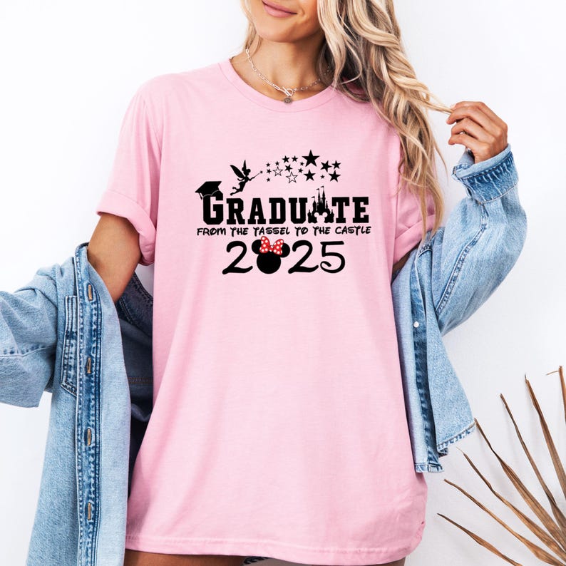 以下が含まれることがあります： ピンクのTシャツに、黒いグラフィックで「Graduate from the tassel to the castle 2025」と書いてあり、シンデレラ城の黒いシルエットとティンカーベルのグラフィックがあります。グラフィックには、赤いリボンが付いた黒いミッキーマウスの頭も含まれています。