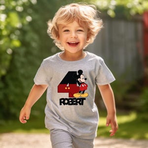 Puede incluir: Una camiseta gris con un gr&aacute;fico de Mickey Mouse y el n&uacute;mero 4. El nombre "ROBERT" est&aacute; impreso debajo. La camiseta la lleva un ni&ntilde;o peque&ntilde;o.