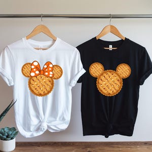 K&ouml;nnte beinhalten: Zwei T-Shirts, eines wei&szlig; und eines schwarz, jeweils mit einem Design zum Thema Kuchen. Das wei&szlig;e T-Shirt hat ein kuchenf&ouml;rmiges Design mit einer Schleife, w&auml;hrend das schwarze T-Shirt ein Kuchendesign hat. Beide h&auml;ngen an Holzb&uuml;geln.