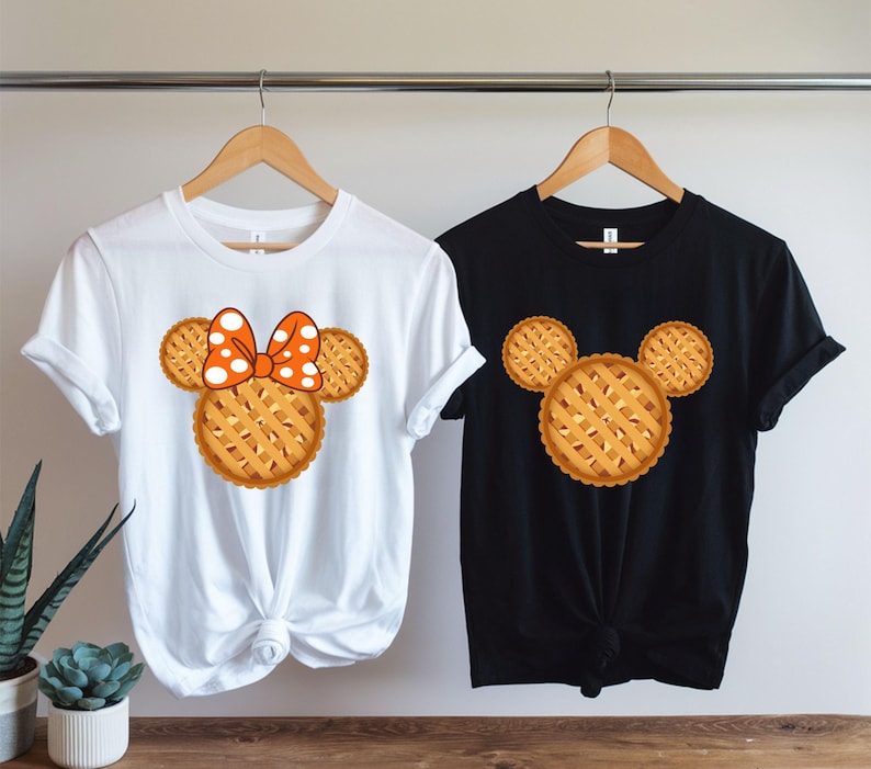 K&ouml;nnte beinhalten: Zwei T-Shirts, eines wei&szlig; und eines schwarz, jeweils mit einem Design zum Thema Kuchen. Das wei&szlig;e T-Shirt hat ein kuchenf&ouml;rmiges Design mit einer Schleife, w&auml;hrend das schwarze T-Shirt ein Kuchendesign hat. Beide h&auml;ngen an Holzb&uuml;geln.