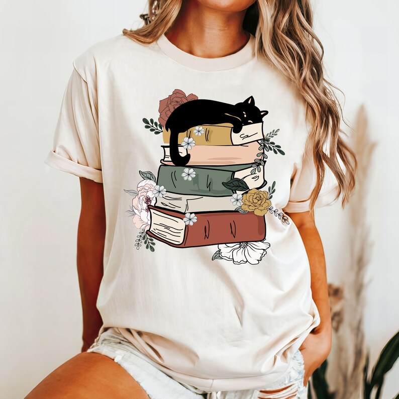 May include: A beige t-shirt with a graphic of a black cat sleeping on a stack of books with floral accents.