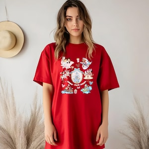 May include: A red t-shirt featuring a colorful graphic design with cartoon characters and a castle. The design includes Mickey and Minnie Mouse, a teacup, and other whimsical elements. The shirt is a casual, oversized style.
