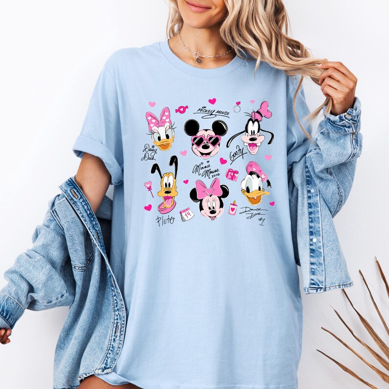 May include: Light blue t-shirt featuring a collage of Disney characters including Mickey Mouse, Minnie Mouse, Donald Duck, and Goofy. The design incorporates pink hearts and character signatures. The shirt is a relaxed fit.