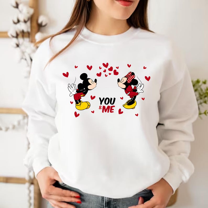 May include: White sweatshirt with a graphic print of Mickey Mouse and Minnie Mouse kissing. The text "YOU & ME" is printed below the characters. The characters are surrounded by red hearts.
