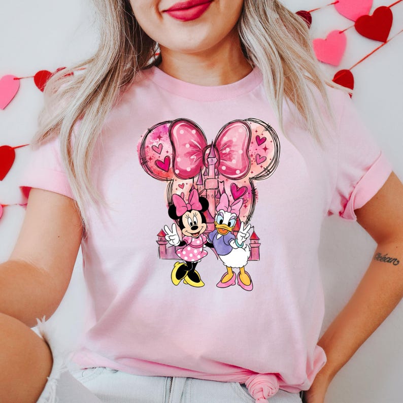 May include: Pink t-shirt featuring a Minnie Mouse and Daisy Duck graphic. The design includes a large pink bow, hearts, and a castle. Minnie Mouse is wearing a pink dress with white polka dots, and Daisy Duck is in a purple top.