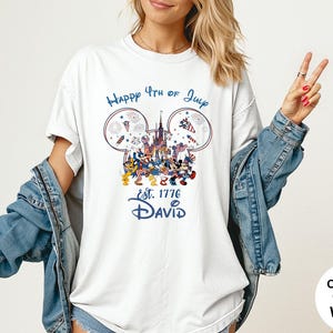 May include: White t-shirt with a patriotic design featuring Mickey Mouse ears, fireworks, and a castle. The text reads "Happy 4th of July" and "Est. 1776 David" in blue. The shirt is a comfort color.