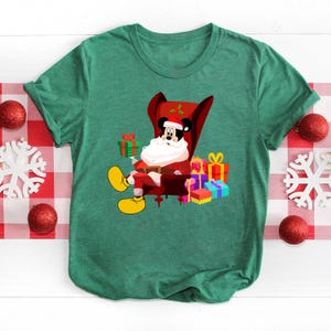 Op de afbeelding: Groen t-shirt met een cartoon Mickey Mouse verkleed als kerstman, zittend in een rode fauteuil en een groen cadeau vasthoudend. Cadeaus in verschillende kleuren zijn rond de stoel gestapeld. Het shirt heeft een ronde hals en korte mouwen.