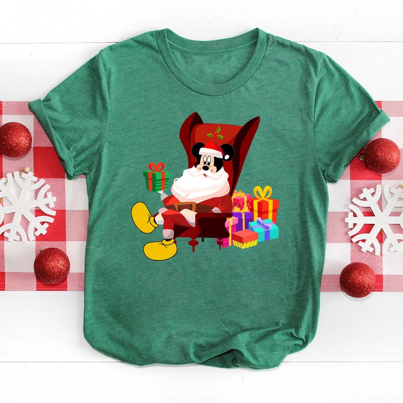 Op de afbeelding: Groen t-shirt met een cartoon Mickey Mouse verkleed als kerstman, zittend in een rode fauteuil en een groen cadeau vasthoudend. Cadeaus in verschillende kleuren zijn rond de stoel gestapeld. Het shirt heeft een ronde hals en korte mouwen.