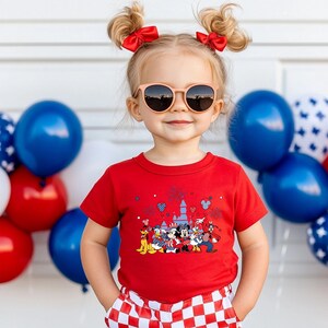 K&ouml;nnte beinhalten: Ein junges Kind tr&auml;gt ein rotes T-Shirt mit einem Disney-Charakter-Motiv. Das Kind tr&auml;gt auch eine Sonnenbrille und hat zwei Z&ouml;pfe mit roten Schleifen. Blaue und wei&szlig;e Luftballons sind im Hintergrund.