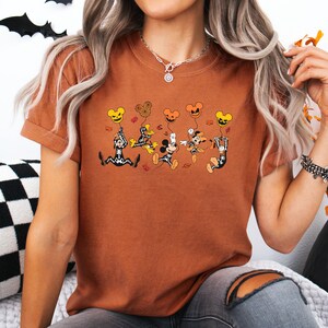 May include: A rust-coloured t-shirt with a Halloween cartoon design. The graphic shows cartoon characters, including Mickey Mouse, in Halloween costumes, holding pumpkin and Mickey Mouse-shaped balloons. The design uses black, orange, and yellow.