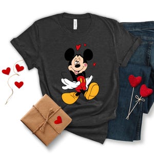 May include: Dark gray t-shirt featuring a smiling Mickey Mouse graphic with red hearts. The cartoon character is wearing a red shirt, white gloves, and yellow shoes. The shirt is paired with blue jeans and a wrapped gift.