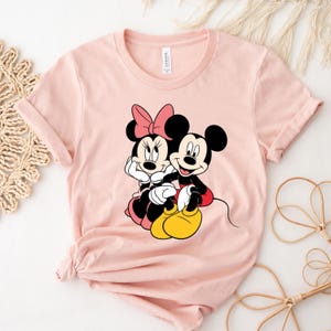 May include: A pink t-shirt with a graphic print of Mickey Mouse and Minnie Mouse sitting together. The characters are in their signature red and black colors.