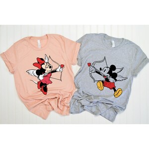 May include: Two t-shirts with a Valentine's Day theme. One shirt is peach coloured with a graphic of Minnie Mouse dressed as Cupid. The other shirt is grey with a graphic of Mickey Mouse dressed as Cupid.