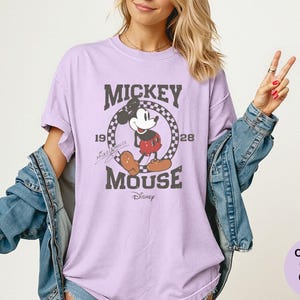 May include: A light purple t-shirt featuring a distressed graphic of Mickey Mouse. The design includes the text "Mickey Mouse" and the years "1928" and "1928" within a circular checkered border. The Disney logo is at the bottom.