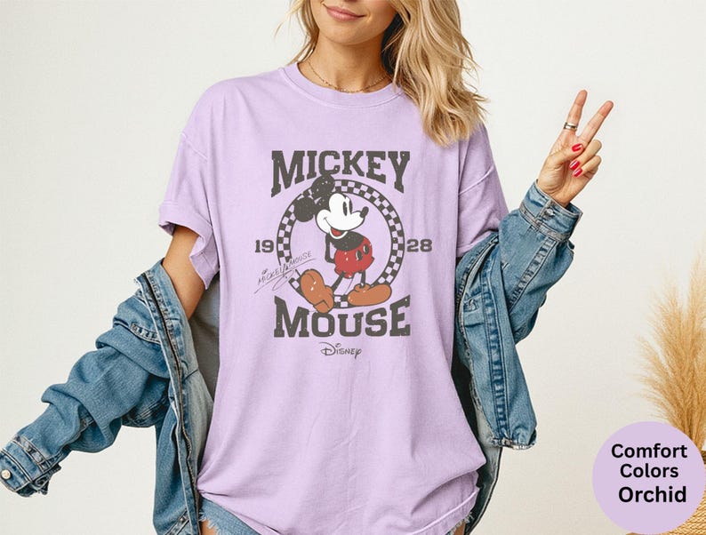 May include: A light purple t-shirt featuring a distressed graphic of Mickey Mouse. The design includes the text "Mickey Mouse" and the years "1928" and "1928" within a circular checkered border. The Disney logo is at the bottom.