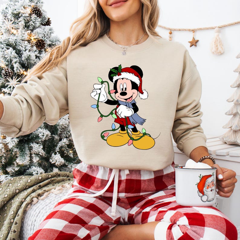 Puede incluir: Sudadera beige con un gr&aacute;fico festivo de Mickey Mouse. Mickey lleva un gorro y una bufanda de Pap&aacute; Noel, sosteniendo luces navide&ntilde;as. La imagen tambi&eacute;n muestra un pantal&oacute;n de pijama a cuadros rojos y blancos y una taza con un dise&ntilde;o de Pap&aacute; Noel.