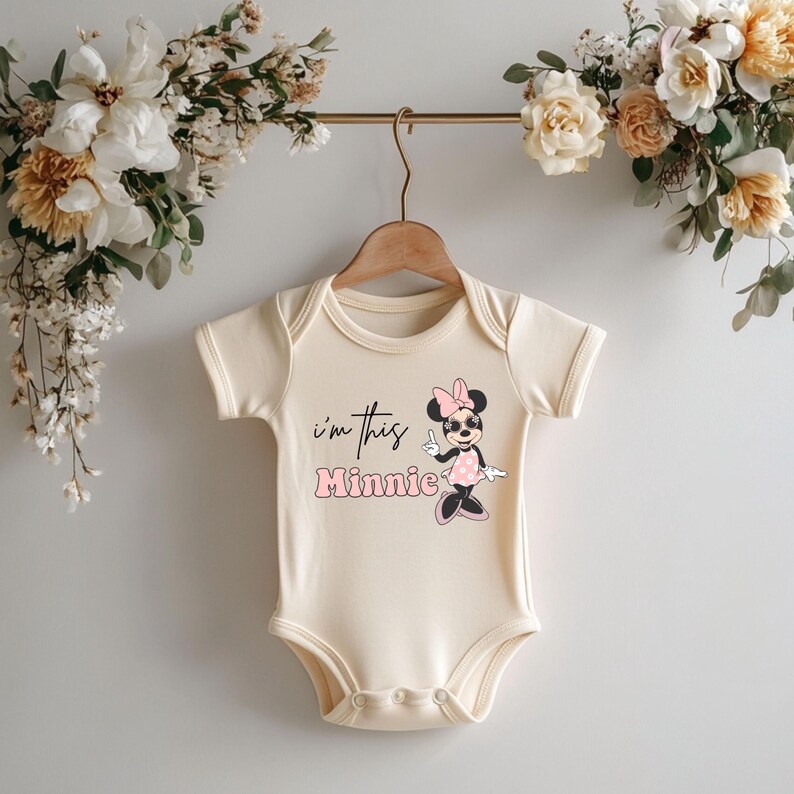 Pu&ograve; includere: Body per neonati color crema con maniche corte, appeso a un gancio di legno. Il body presenta la scritta "I'm this Minnie" e una grafica di Minnie Mouse in rosa, nero e bianco. Composizione floreale sullo sfondo.