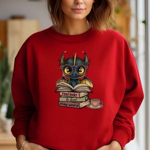 May include: Red sweatshirt with a cute cartoon dragon wearing glasses and reading a book. The text on the sweatshirt says "read books be kind stay weird".