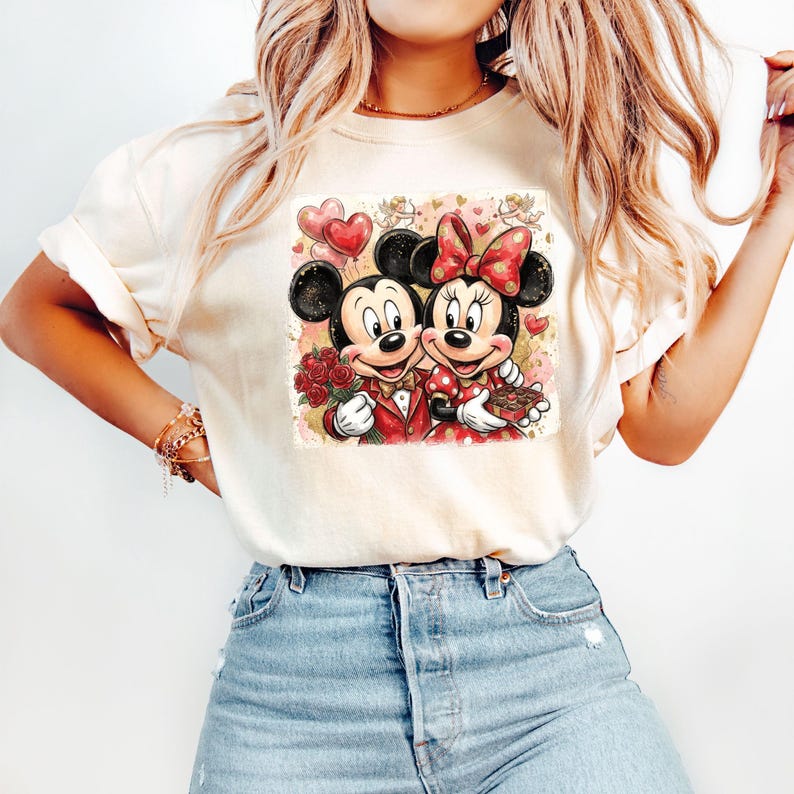 May include: Cream-colored t-shirt featuring a graphic of Mickey and Minnie Mouse in formal attire, holding roses and a gift. Minnie wears a polka-dot dress and bow. Mickey holds a heart-shaped balloon. The design has a watercolor effect.