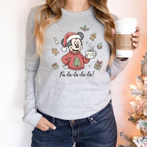 May include: A gray long-sleeve shirt featuring a Christmas-themed Mickey Mouse graphic. Mickey is wearing a Santa hat and a red sweater with a Christmas tree. The shirt has the text "Fa-la-la-la-la!" below the graphic.