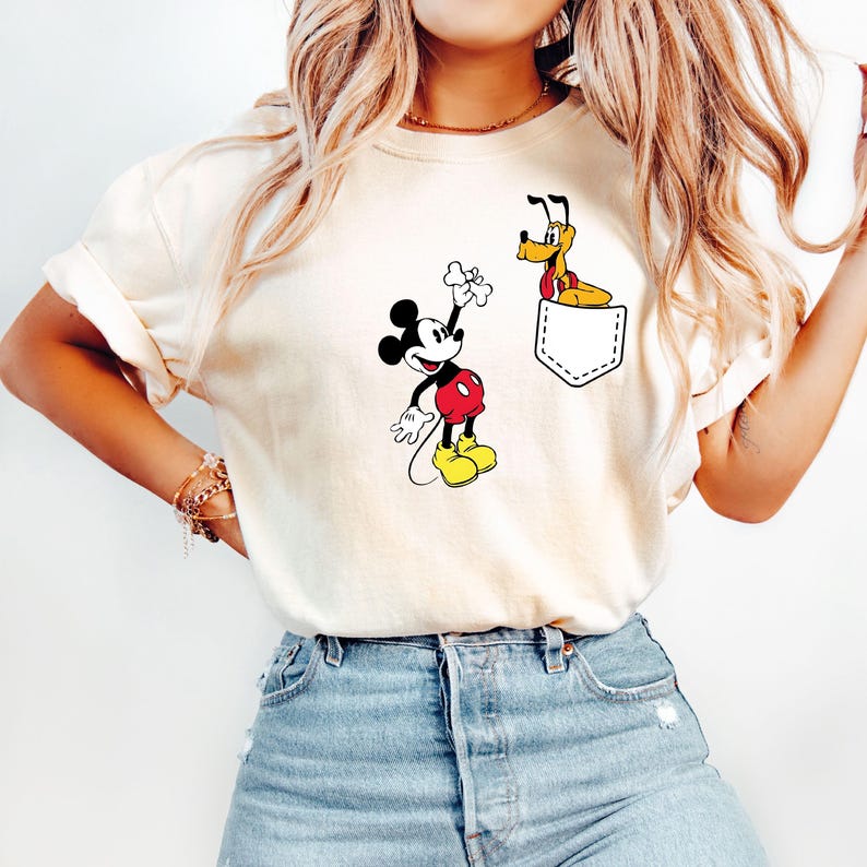 Puede incluir: Una camiseta blanca con un bolsillo que presenta una imagen de dibujos animados de Mickey Mouse y Pluto. Mickey Mouse sostiene un hueso y Pluto asoma por el bolsillo.