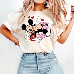 May include: A white t-shirt featuring a graphic of Mickey Mouse and Minnie Mouse kissing. The graphic is in black, red, and pink. There are small red hearts around the graphic.