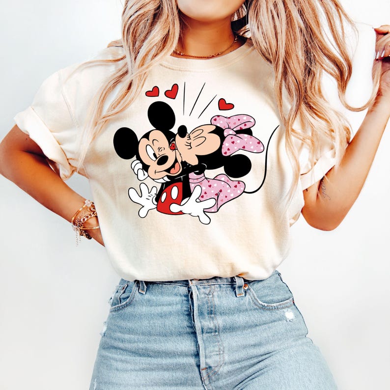 May include: A white t-shirt featuring a graphic of Mickey Mouse and Minnie Mouse kissing. The graphic is in black, red, and pink. There are small red hearts around the graphic.