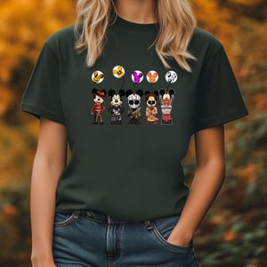 May include: Dark green t-shirt featuring a graphic of six cartoon characters dressed as horror movie icons. Each character holds a balloon with a Mickey Mouse head design. The balloons are yellow, orange, pink, and white.
