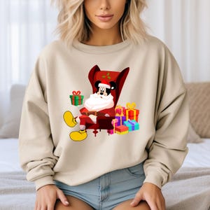 Op de afbeelding: Beige sweatshirt met een cartoon Mickey Mouse verkleed als kerstman, zittend in een rode stoel. Mickey is omringd door kleurrijk verpakte cadeaus. Het ontwerp is feestelijk en geschikt voor de feestdagen.