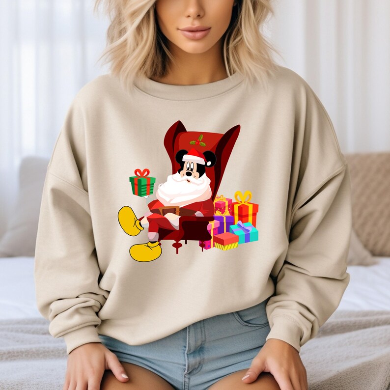 Op de afbeelding: Beige sweatshirt met een cartoon Mickey Mouse verkleed als kerstman, zittend in een rode stoel. Mickey is omringd door kleurrijk verpakte cadeaus. Het ontwerp is feestelijk en geschikt voor de feestdagen.