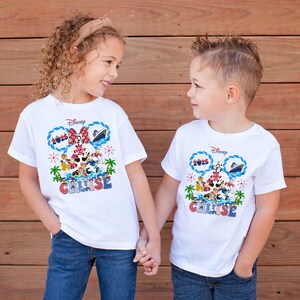 May include: Two children wearing white t-shirts with a Disney Cruise design. The shirts feature Mickey and Minnie Mouse, a cruise ship, and the word "CRUISE" in blue and red. The children are holding hands.