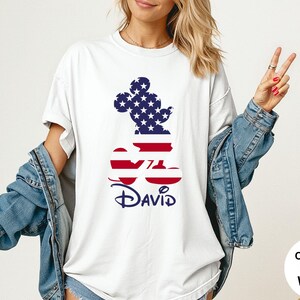May include: White t-shirt featuring a Mickey Mouse silhouette filled with a US flag design. The name "David" is printed in navy blue below the design. The shirt is part of the Comfort Colors collection.