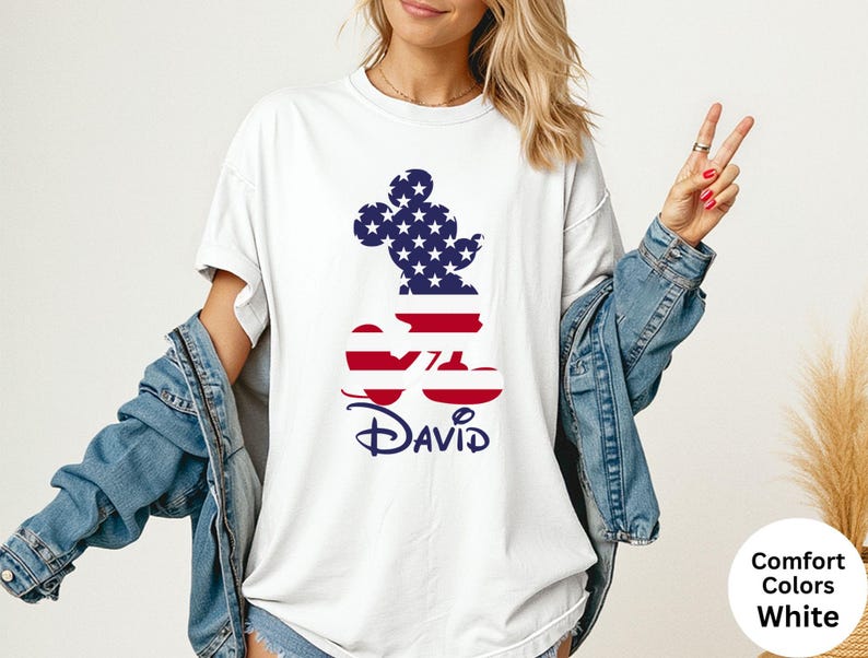 May include: White t-shirt featuring a Mickey Mouse silhouette filled with a US flag design. The name "David" is printed in navy blue below the design. The shirt is part of the Comfort Colors collection.