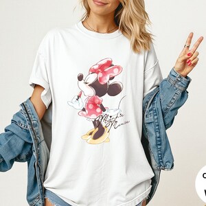 K&ouml;nnte beinhalten: Wei&szlig;es T-Shirt mit Minnie Mouse-Grafik. Das Design zeigt Minnie mit roter Schleife und gepunktetem Kleid, mit dem Schriftzug "Minnie Mouse" in Schreibschrift. Teil der Comfort Colors-Kollektion.