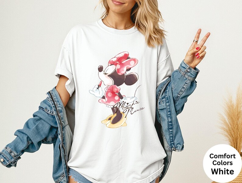 K&ouml;nnte beinhalten: Wei&szlig;es T-Shirt mit Minnie Mouse-Grafik. Das Design zeigt Minnie mit roter Schleife und gepunktetem Kleid, mit dem Schriftzug "Minnie Mouse" in Schreibschrift. Teil der Comfort Colors-Kollektion.