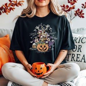 May include: Black t-shirt featuring a Halloween-themed graphic with Disney characters like Mickey Mouse, Donald Duck, and Goofy. The design includes a carved pumpkin and bats. The person is holding an orange jack-o'-lantern mug.