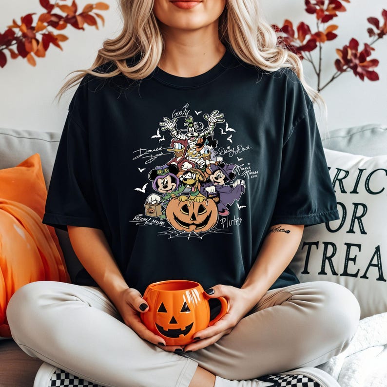 May include: Black t-shirt featuring a Halloween-themed graphic with Disney characters like Mickey Mouse, Donald Duck, and Goofy. The design includes a carved pumpkin and bats. The person is holding an orange jack-o'-lantern mug.