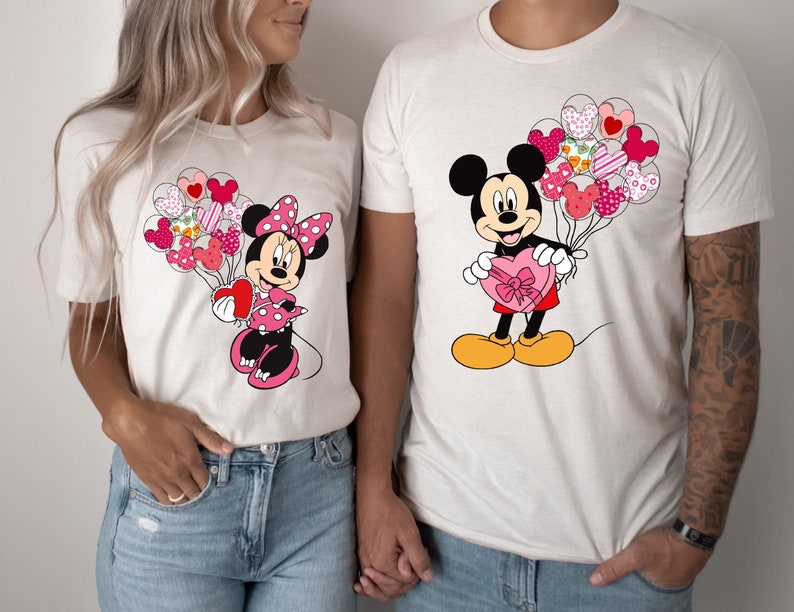 May include: A white t-shirt with a pink and red Mickey Mouse holding a heart-shaped box of chocolates. The Mickey Mouse is surrounded by pink and red heart-shaped balloons.