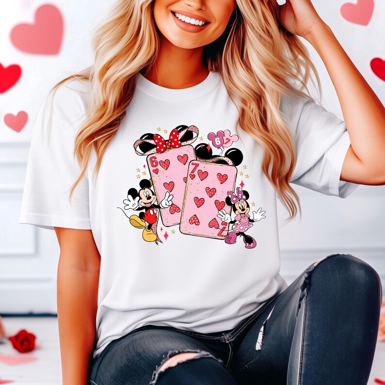 May include: White t-shirt featuring a graphic of Mickey and Minnie Mouse with playing cards. The cards are pink with red hearts and the numbers 6 and 7. Mickey and Minnie are in their classic outfits. A red bow and heart balloons are also featured.