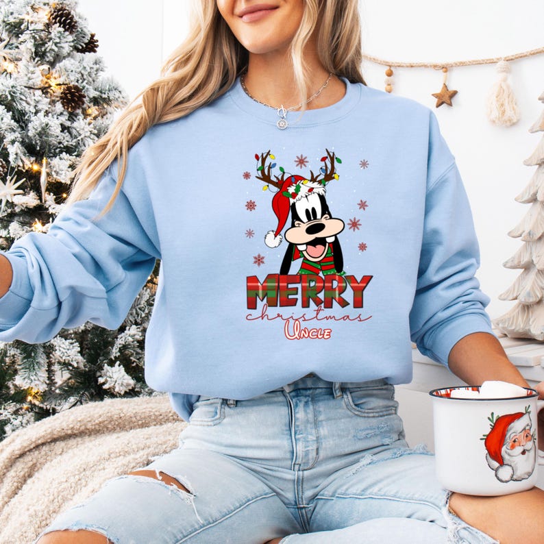 May include: Light blue sweatshirt featuring a cartoon character with reindeer antlers, a Santa hat, and a scarf. The text "MERRY Christmas Uncle" is printed in red and white. The sweatshirt is a festive holiday design.