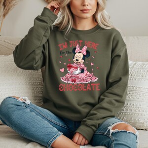 May include: An olive green sweatshirt featuring a graphic of Minnie Mouse holding a heart-shaped gift box. The text "I'M JUST HERE for the CHOCOLATE" is printed in pink and red. The design includes pink hearts and a pink polka dot bow.