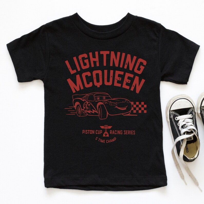 May include: Black short-sleeved t-shirt with a red graphic of Lightning McQueen from the Cars movie. The graphic includes the text "Lightning McQueen", "Piston Cup Racing Series", and "5-Time Champ".