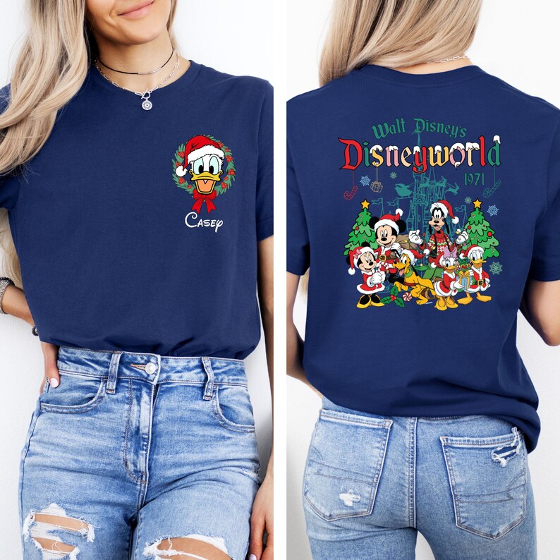 Op de afbeelding: Marineblauw T-shirt met Donald Duck in een kerstkrans en de naam "Casey". De achterkant van het shirt toont "Walt Disney's Disneyworld 1971" met Mickey, Minnie, Goofy, Donald en andere personages in feestelijke kleding.