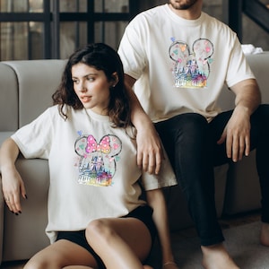 Op de afbeelding: Een stel draagt ​​bijpassende witte T-shirts met een kleurrijke illustratie van een kasteel en Mickey Mouse-oren. Het shirt van de vrouw heeft een roze strik op de oren, terwijl het shirt van de man een eenvoudige omtrek heeft.