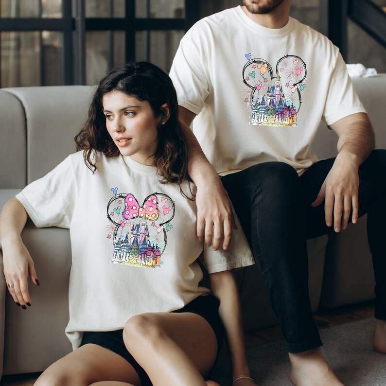 Op de afbeelding: Een stel draagt ​​bijpassende witte T-shirts met een kleurrijke illustratie van een kasteel en Mickey Mouse-oren. Het shirt van de vrouw heeft een roze strik op de oren, terwijl het shirt van de man een eenvoudige omtrek heeft.