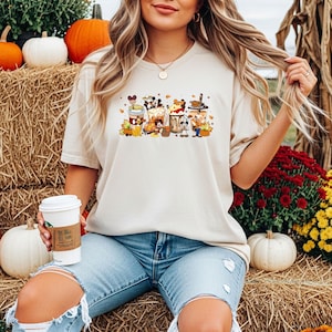 May include: Beige t-shirt featuring a colorful graphic of cartoon coffee cups with fall-themed decorations. The design includes pumpkins, leaves, and character-themed elements. The shirt is paired with light blue jeans.