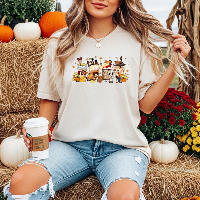May include: Beige t-shirt featuring a colorful graphic of cartoon coffee cups with fall-themed decorations. The design includes pumpkins, leaves, and character-themed elements. The shirt is paired with light blue jeans.
