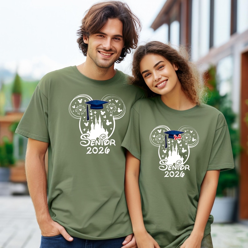 Puede incluir: Dos camisetas verde oliva con un dise&ntilde;o de graduaci&oacute;n. El dise&ntilde;o presenta un contorno blanco de las orejas de Mickey Mouse, un birrete, un castillo y el texto "Senior 2026". Una camiseta tiene una gorra azul, la otra un lazo rojo.