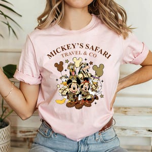 K&ouml;nnte beinhalten: Rosa T-Shirt mit einem Aufdruck von Mickey Mouse und seinen Freunden in Safari-Outfits. Der Text "Mickey's Safari Travel & Co" ist auf dem Shirt aufgedruckt.